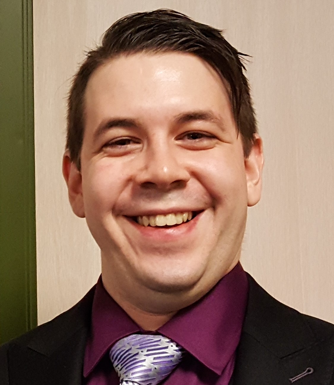 A picture of Eric De Wildt wearing a black suit, deep purple shirt, and silver and purple tie. Eric has long bangs and his hair is combed to his left and held in place with product. Eric is smiling and is a bit chubby.